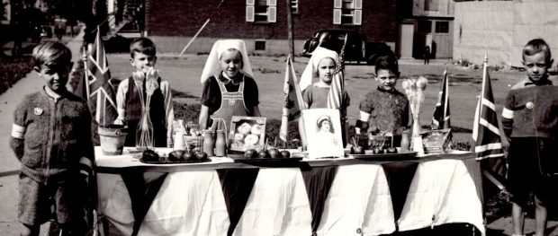 Portrait of six children, two girls in the middle and two boys on each side, behind a table with food, four Union Jacks and two vases of flowers. The young girls are wearing nun-like- head coverings and the boys are sporting lapel badges on their vests.