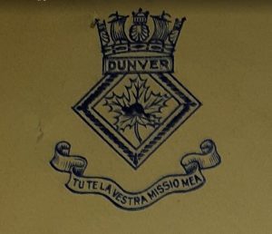 Emblème du HMCS Dunver imprimé en noir sur papier beige. C’est un losange avec un castor sur une feuille d’érable à l’intérieur. Une couronne superpose le haut du losange. En dessous, une inscription sur un ruban.