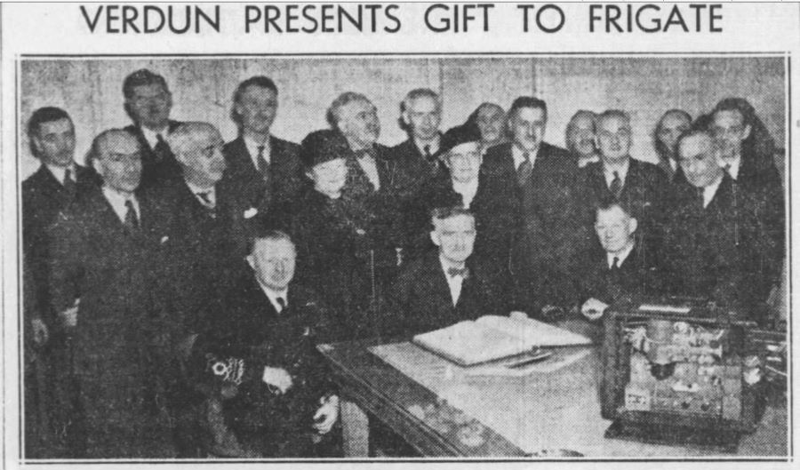 Black and white photograph titled “Verdun presents gift to frigate.” In a room, at the end of a table with a projector and an open ledger on it, three people are sitting and 16 standing.