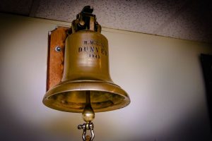 Colour photograph of a bell hanging on a wall in a room. The inscription HMCS DUNVER 1943 is engraved on the golden bell.