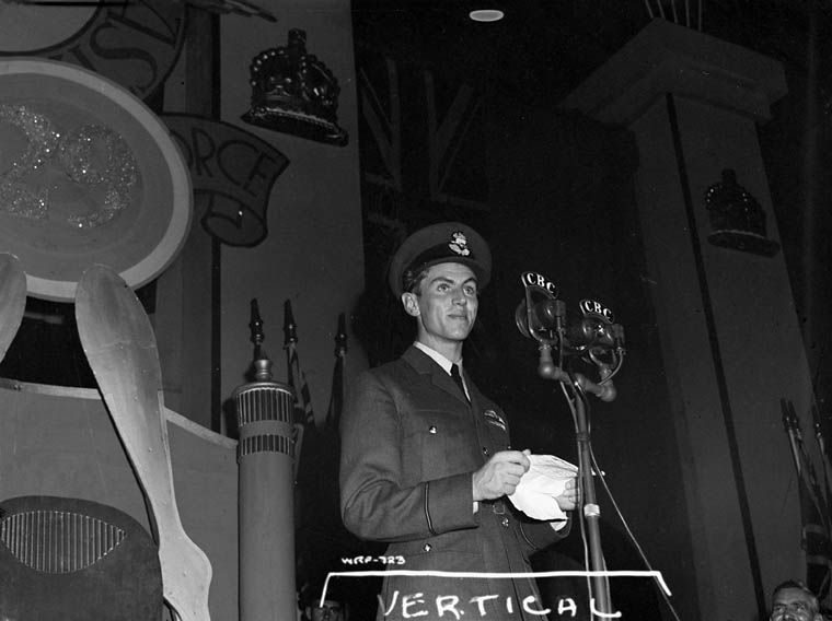 Black and white photograph of a young man in uniform delivering a speech and holding a crumpled piece of paper. Behind him, a plane propeller, the number 29 in a circular frame and a Union Jack. In the foreground, CBC microphones.