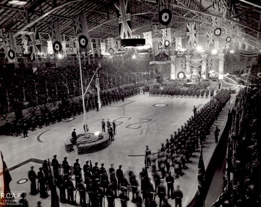 Black and white photograph. A stage is set up at the end of a skating rink in an arena. Rows of servicemen in front and on the sides. Aviation flags and the Union Jack hang from the ceiling. The bleachers are filled with spectators.