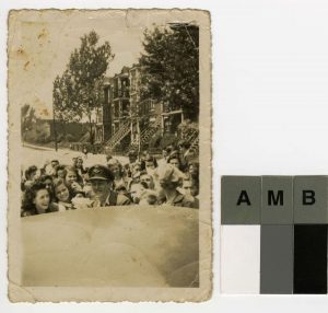 Black and white photograph of a young man in uniform, smiling as he is getting into a car. He is surrounded by about 20 young people, both boys and girls. In the background, a street, buildings and trees.