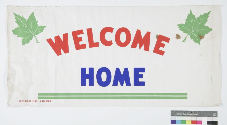 Rectangular cotton banner with colour paint. A green maple leaf in each upper corner, and below, in a semi-circle the word “Welcome” in red, in the centre the word “Home” in blue. At the bottom, two parallel green lines.