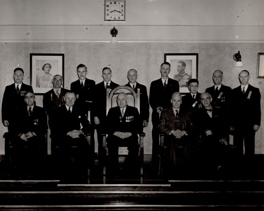 Photographie noir et blanc sur laquelle se trouvent cinq hommes assis et neuf hommes debout derrière.  Les portraits du roi George VI et de la Reine Elizabeth sont accrochés sur le mur en arrière-plan. Une horloge indique 8 h 18.