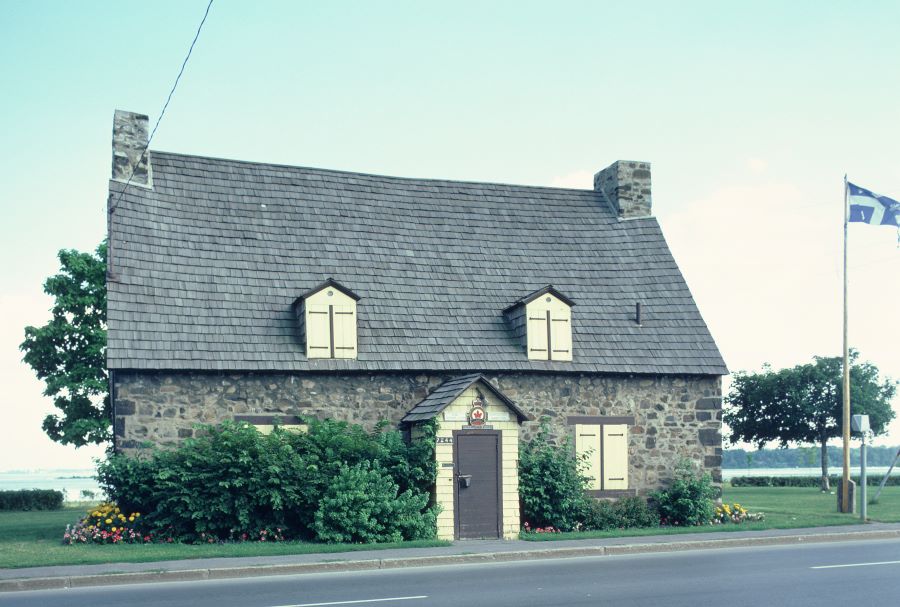 Colour photograph of an old Breton-style house with an entrance door at the centre, two windows on the ground floor and two dormers in the attic. The Canadian Legion emblem is featured above the door. A Québec flag flies to the right of the house. The St. Lawrence River is visible in the background.