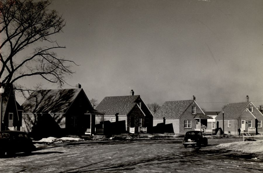 Black and white photograph of four small cottage-style houses along a street. Two cars parked on the street are visible in the foreground. To the left, half of a big tree.