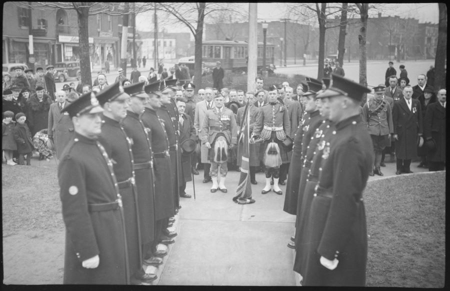 Black and white photograph. In the centre, 12 servicemen in uniform form an honour guard before two servicemen in kilts flanking a man in civilian clothes , holding a flag. Behind them are some 50 dignitaries and spectators. In the background, a few trees, the street and buildings.