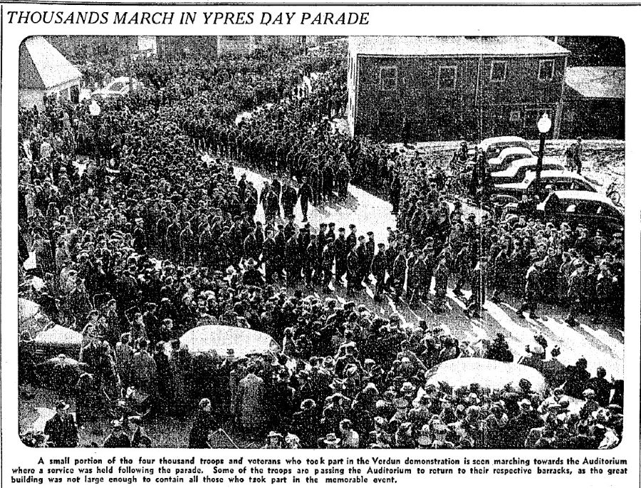 Black and white newspaper article titled “Thousands March in Ypres Day Parade.” Below, a large photograph of hundreds of people marching in the street, past parked cars surrounded by onlookers, followed by a 65-word text.