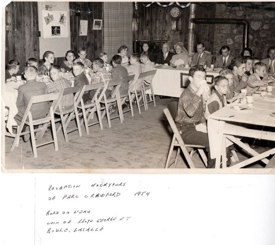 Black and white photograph of seven adults seated at a table. Two groups of children are seated around two other tables, 14 of the children are to the left and seven others to right of the table where the adults are seated. In the background, a stone wall, a clock and framed pictures.