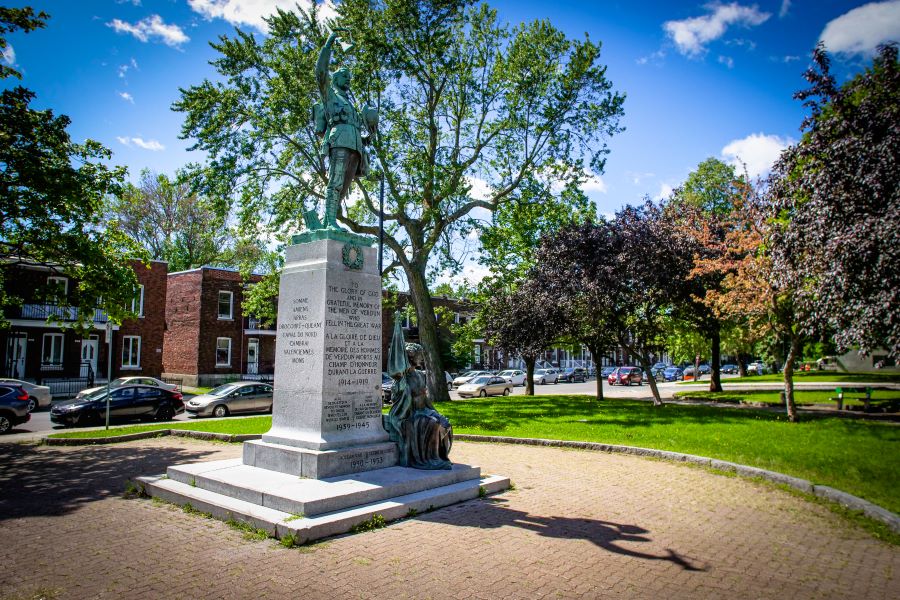 Colour photograph of a memorial comprised of two green-tinted statues. One statue is of a soldier and is placed on a grey pedestal, the other statue is of a woman, placed at the foot of the memorial. In the background, trees, cars parked in the street and brick buildings.