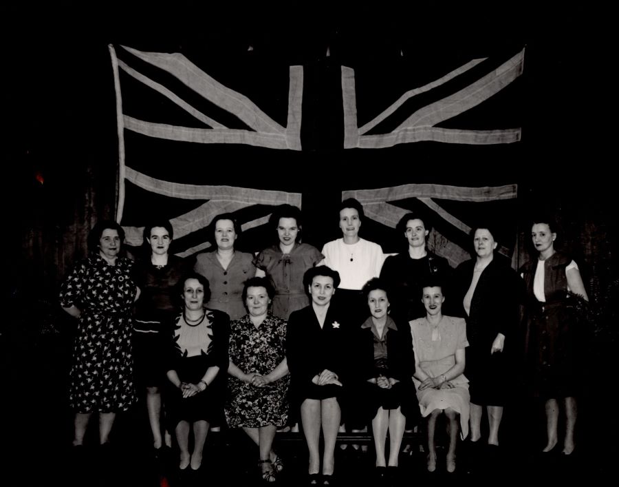Photographie en noir et blanc d'un groupe de treize femmes devant un grand drapeau britannique. Elles sont placées en deux rangées.