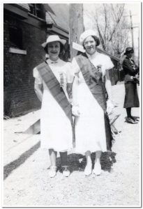 Black and white photograph. In the foreground, two women standing in the street dressed in uniform: a light-coloured dress with a chequered scarf worn as a bandoleir. In the background, a woman, a building and a tree.