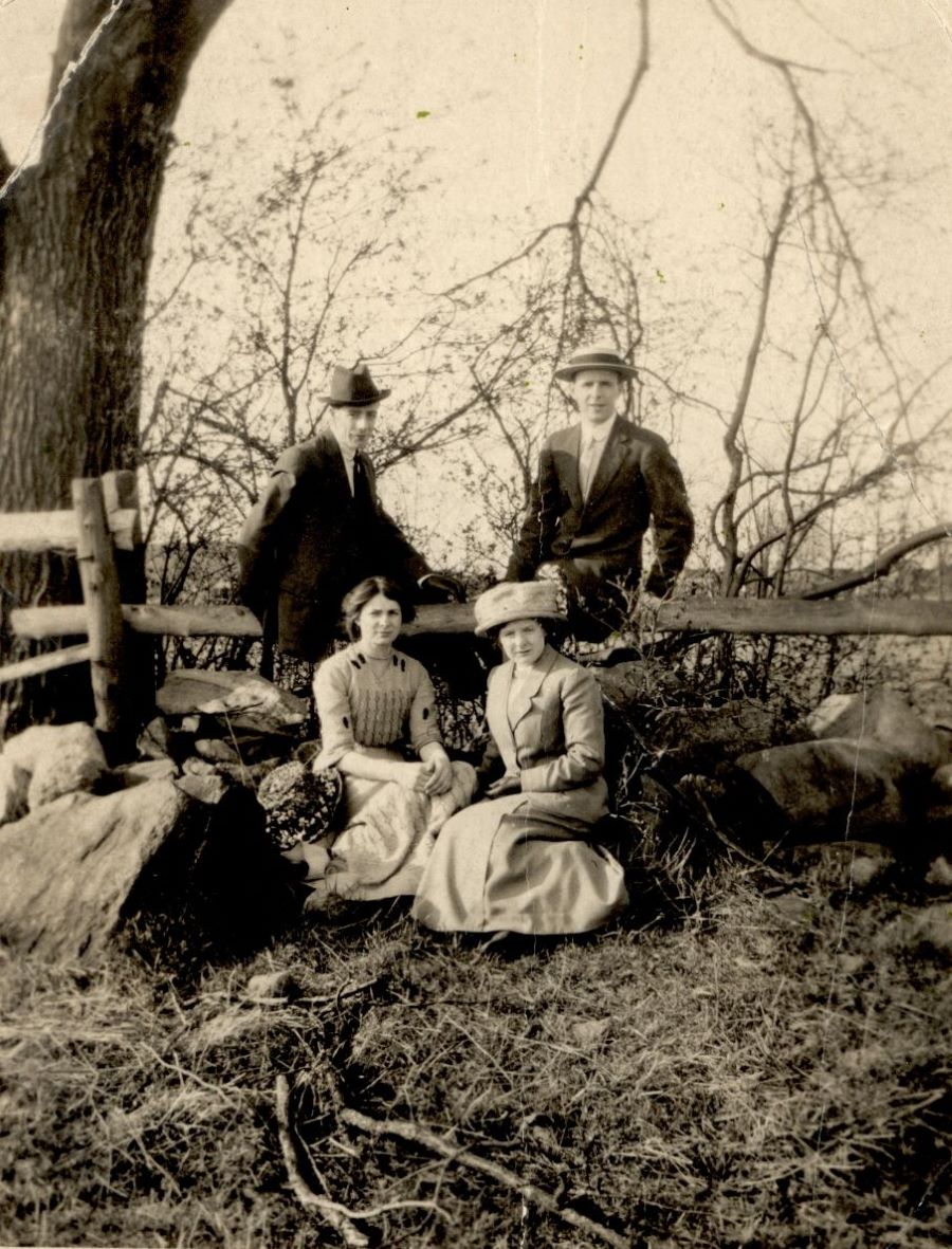 Black and white photograph of four elegantly dressed adults: two men standing against a wooden farm fence, with two women sitting on the grass in front of them. In the background, a tree and a field.