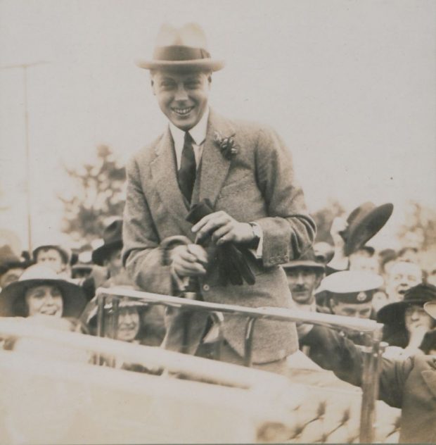 Black and white photograph of the Prince of Wales, wearing a suit. Standing, the prince smiles at the camera, with a crowd in the background welcoming him.