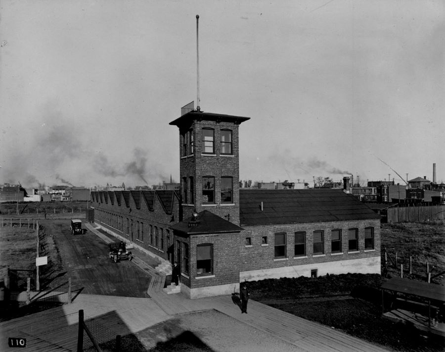 Black and white photograph of an industrial building with a corner tower, several windows on the facades and a saw-toothed roof. In the background, smoke billowing out of industrial stacks.