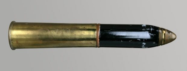 Colour photograph of a brass shell with a portion near the tip painted black. It is 57 cm in length, with an outside diameter of 13 cm.