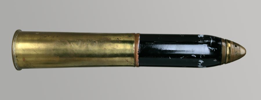 Photographie en couleurs d'un obus doré fabriqué en laiton avec une partie peinte en noire près de la pointe. Il a une longueur de 57 cm et un diamètre extérieur de 13 cm.