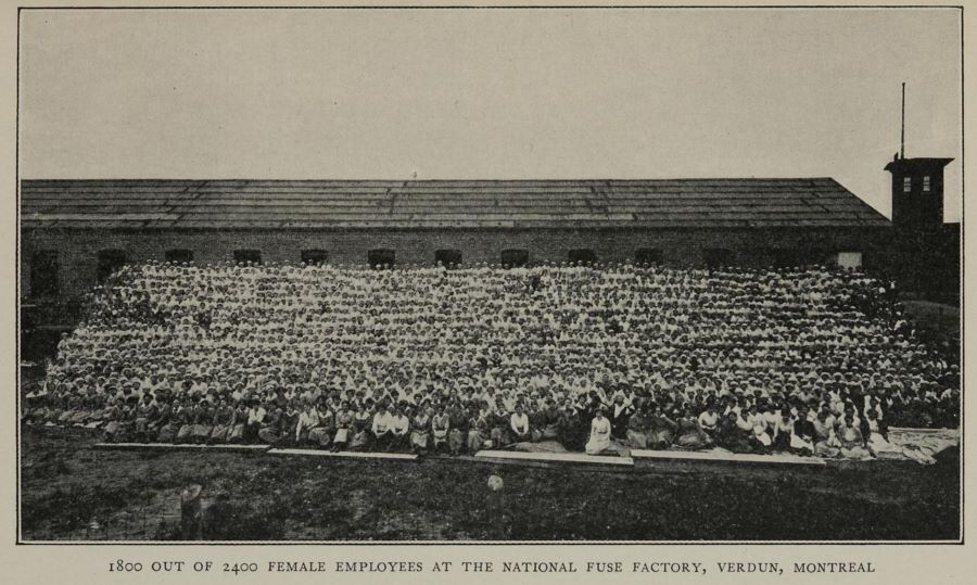 Photographie en noir et blanc montrant 1 800 femmes assises en une vingtaine de rangées sur une petite colline devant l'usine de munitions.