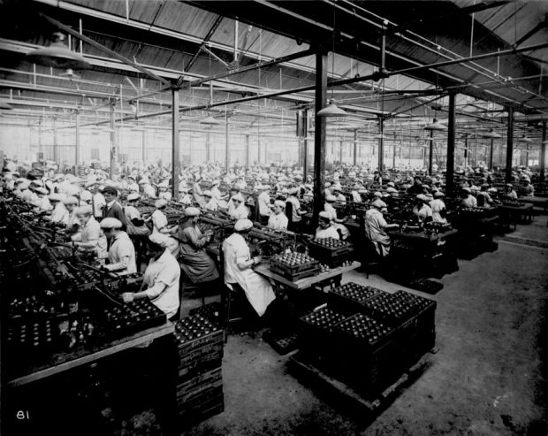 Black and white photograph of several rows of women at work inside the factory. They are seated close together at their work stations. In one of the rows, a man is supervising the work.