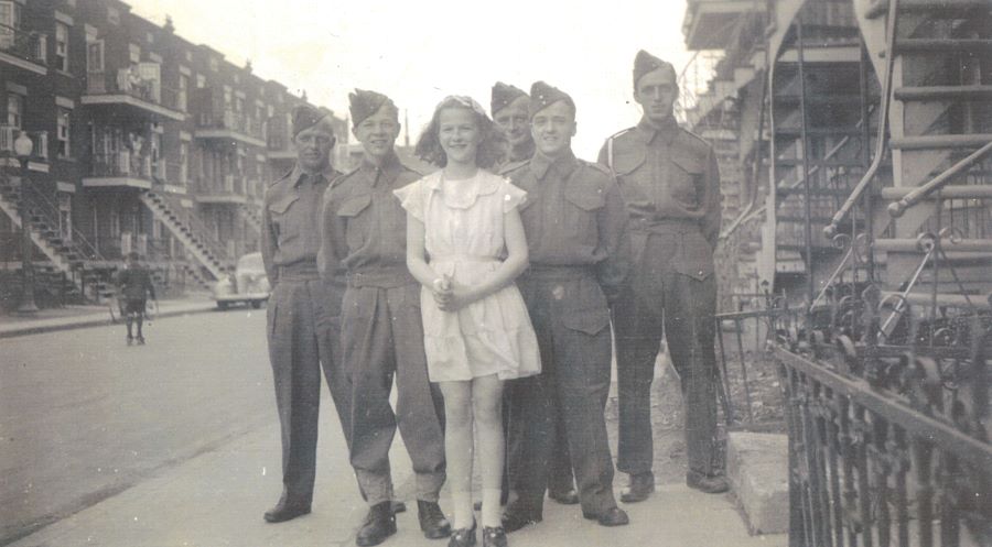 Black and white photograph of six people standing on the sidewalk, a young girl and five servicemen in uniform. In the background, triplexes with staircases and balconies.