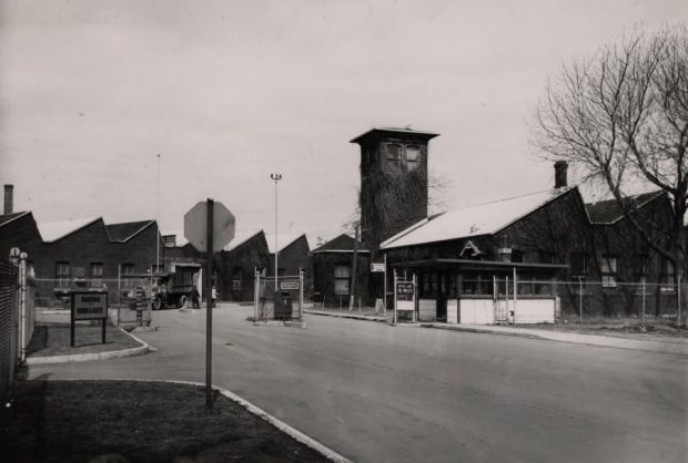 Black and white photograph of the road entrance leading to the facilities of the old munitions factory. A checkpoint is set up at the entrance and the facilities are fenced in.
