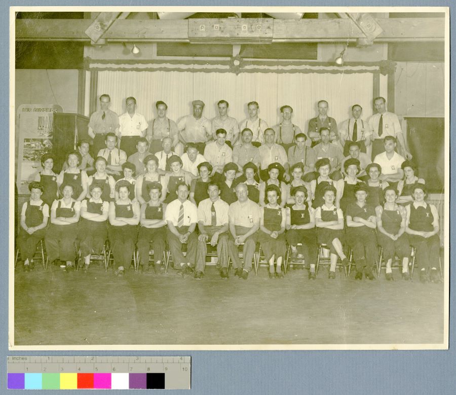 Photographie en noir et blanc de 50 personnes en uniforme de travail à l'intérieur de l'usine. 3 rangées assises et une debout : les deux premières sont majoritairement des femmes et les deux dernières des hommes.