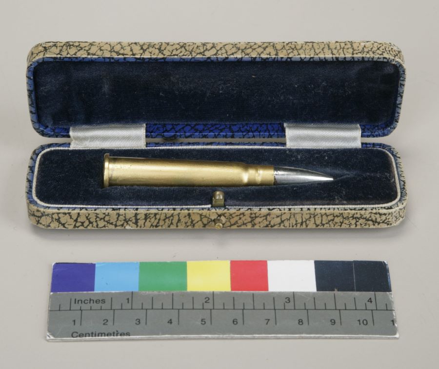 Colour photograph of an open case with a blue velvet interior containing a brass cartridge with a silver tip. The case is 2.5 cm high, 12.1 cm long and 3.5 cm wide.