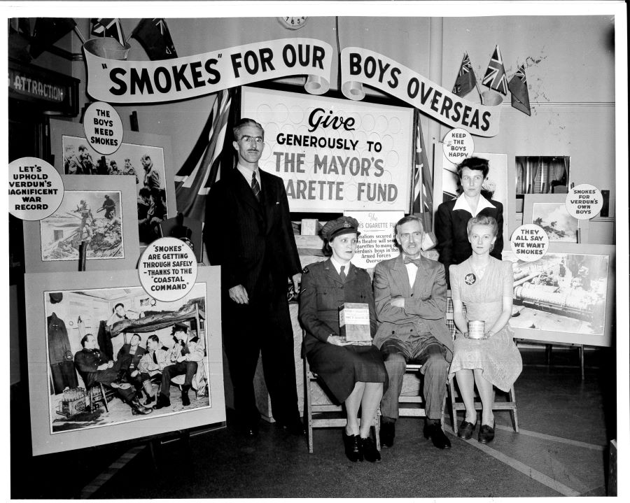 Black and white photograph of two men and three women in front of a promotional display featuring various slogans and several photographs of soldiers in their daily lives.