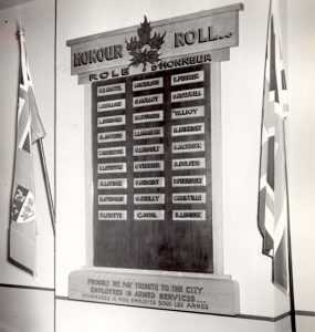 Photographie noir et blanc d’un tableau d’honneur accroché à un mur entre deux drapeaux, un canadien, l’autre britannique. Sur le tableau, 27 petites plaques rectangulaires avec les noms d’employés en service actif. 