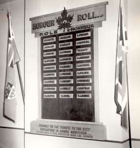 Black and white photograph of an honour roll hung on a wall between two flags, one Canadian and one British. On the roll, 27 small rectangular plaques with the names of City employees in active service.