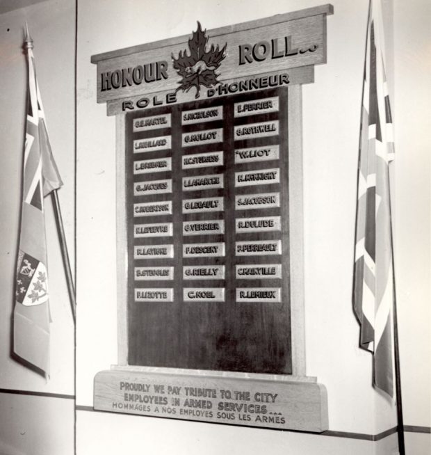 Black and white photograph of an honour roll hung on a wall between two flags, one Canadian and one British. On the roll, 27 small rectangular plaques with the names of City employees in active service.