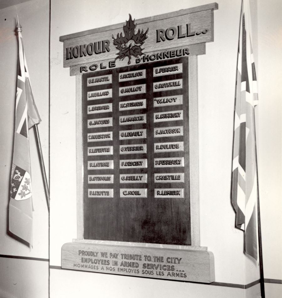 Black and white photograph of an honour roll hung on a wall between two flags, one Canadian and one British. On the roll, 27 small rectangular plaques with the names of City employees in active service.