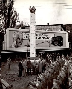Black and white photograph of six people in front of a giant thermometer. Three rows of cadets and several young people watching the scene under grey skies. In the background, two billboards.