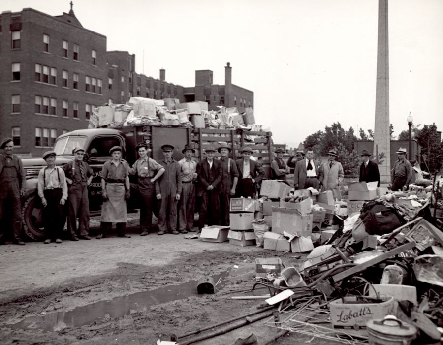 Black and white photograph of 16 men in front of a truck filled with salvaged material. Verdun Hospital is visible in the background and a pile of salvageable material is strewn on the ground in front of the men.