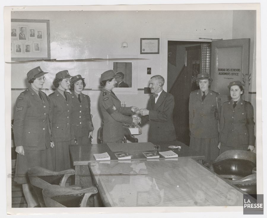 Black and white photograph of a small room with the door open. In the centre, Mayor Wilson handing over the keys to one of the six women in uniform. All seven people are standing around a table.