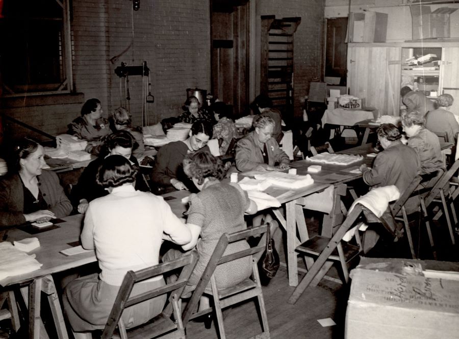 Black and white photograph of 15 women sitting at tables (two long rows) in a room, cutting cloth.