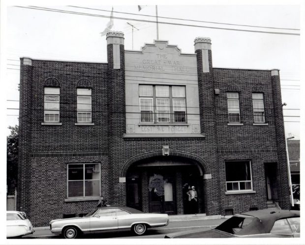 Black and white photograph of a building whose façade features the inscription “The Great War Memorial Hall” and “Lest We Forget.” A man is standing in front of the main door and cars are parked close to the building.