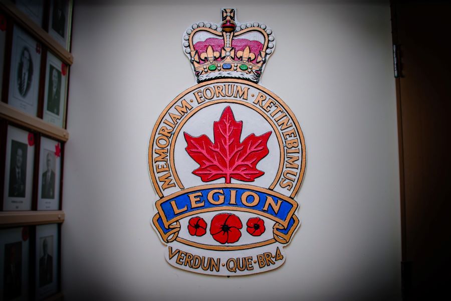 Colour photograph of an emblem on which the word “Legion” is written on a blue ribbon beneath the motto Memoriam Eorum Retinebimus in a half circle framing a red maple leaf and topped by a beige, pink, blue and green crown. Below are three red poppies and the words “Verdun-Que-Br-4.”
