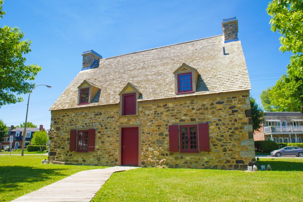 Colour photograph of a stone house with a wooden main door, painted red, two shuttered windows, two dormers and two chimneys. In the background, a street and two buildings.