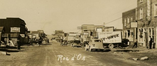 Sepia photograph of an unpaved street lined with buildings on both sides. On the right side, several cars and a truck bearing the inscription St-Onge are visible.