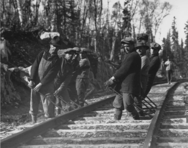 Black and white photograph of a group of eight men with iron bars working on a railway. In the background, a forest.