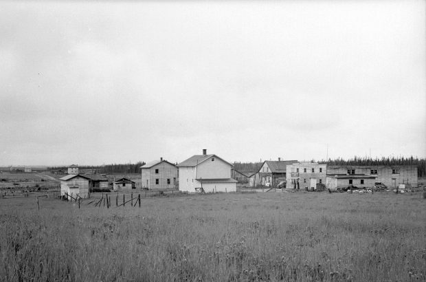 Black and white photograph of a hamlet of about ten buildings, made of logs and planks, surrounded by a field. In the background, a forest.