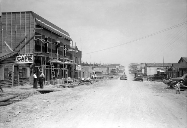 Black and white photograph of a gravel road lined with plank and log buildings. On the left, the Maple Leaf Cafe Chop Suey and Rooms and a building under construction. On the right, several cars and a child walking.
