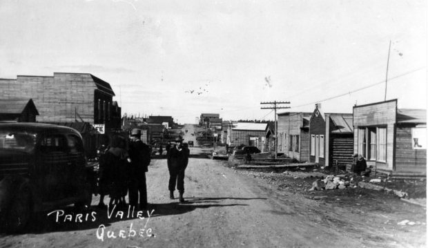 Black and white photograph of a gravel road lined with log cabins and plank buildings. A car and several people are in the middle of the street in the centre of the photo. At the bottom, the inscription “Paris Valley Québec”.