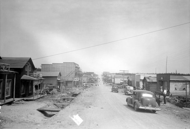 Black and white photograph of a gravel road lined with plant and log buildings. On the right, signs “Paris Cafe” and Royal Restaurant”. In the foreground on the right, the front ends of two cars.