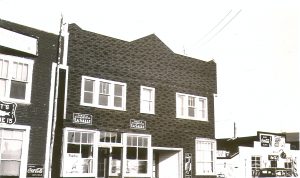 Photographie en noir et blanc d’un immeuble de deux étages recouvert de papier gou-dronné avec une façade de style Boomtown. Deux publicités « Tabac à cigarettes La Salle » sont affichées. À droite, un restaurant et à gauche, un hôtel. 