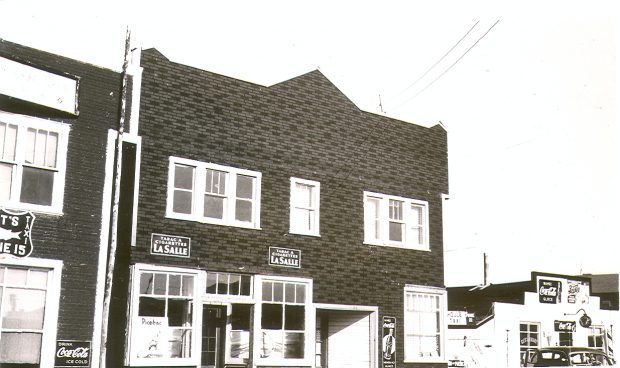 Black and white photograph of a Boomtown-façade two-storey building covered with tar paper. Two La Salle Cigarette Tobacco advertisements are displayed. On the right, a restaurant and on the left, a hotel.