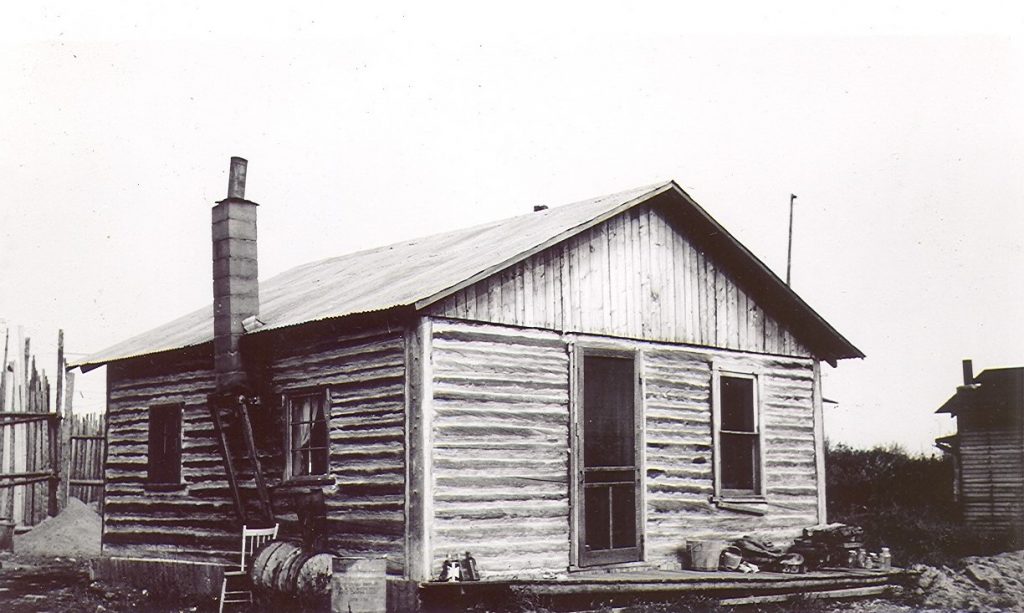 Photographie en noir et blanc d’une cabane en bois rond de construction rudimentaire qui comporte trois fenêtres et une porte. À gauche, une cheminée chambranlante.