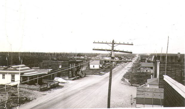 Black and white photograph of a gravel road lined with plank buildings. In the foreground, a pole bearing wires. In the background, a field and a coniferous forest.